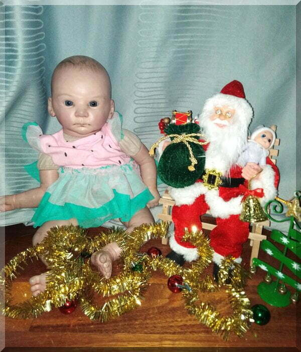 Santa sitting on bench holding a baby elf beside a doll